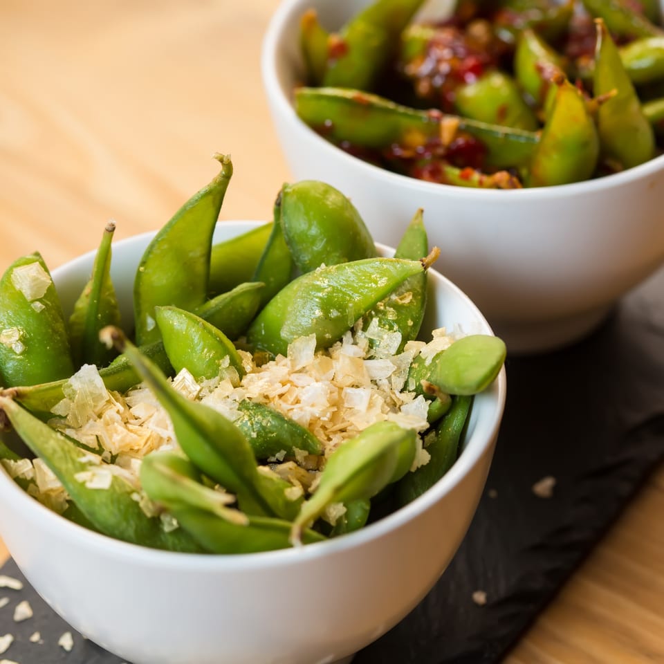 Two bowls of edamame, one with salt flakes and the other with a spicy sauce.