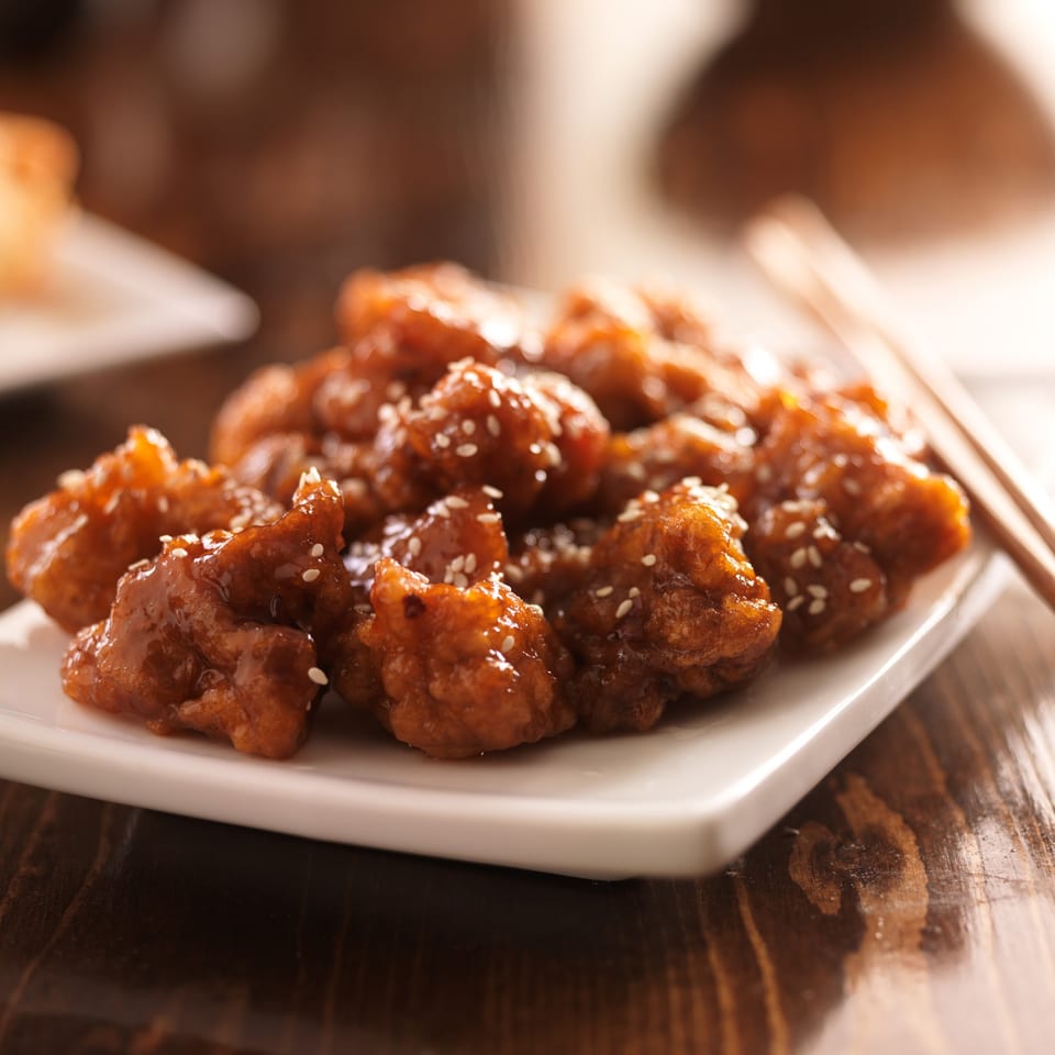 Close-up of Lucky Chopsticks' sesame chicken on a white plate with chopsticks on a wooden table.